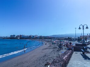 A beach with people swimming and sunbathing, a stone wall, and buildings in the background.