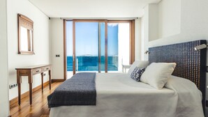 Quarto com cama grande, mesa de cabeceira de madeira, espelho e vista para o mar através de uma porta de correr de vidro.