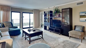 A living room with a sofa, a coffee table, a TV, a bookshelf, and a balcony with curtains. - Destin Beachfront Condo | Stunning Views & Resort Amenities (Destin)