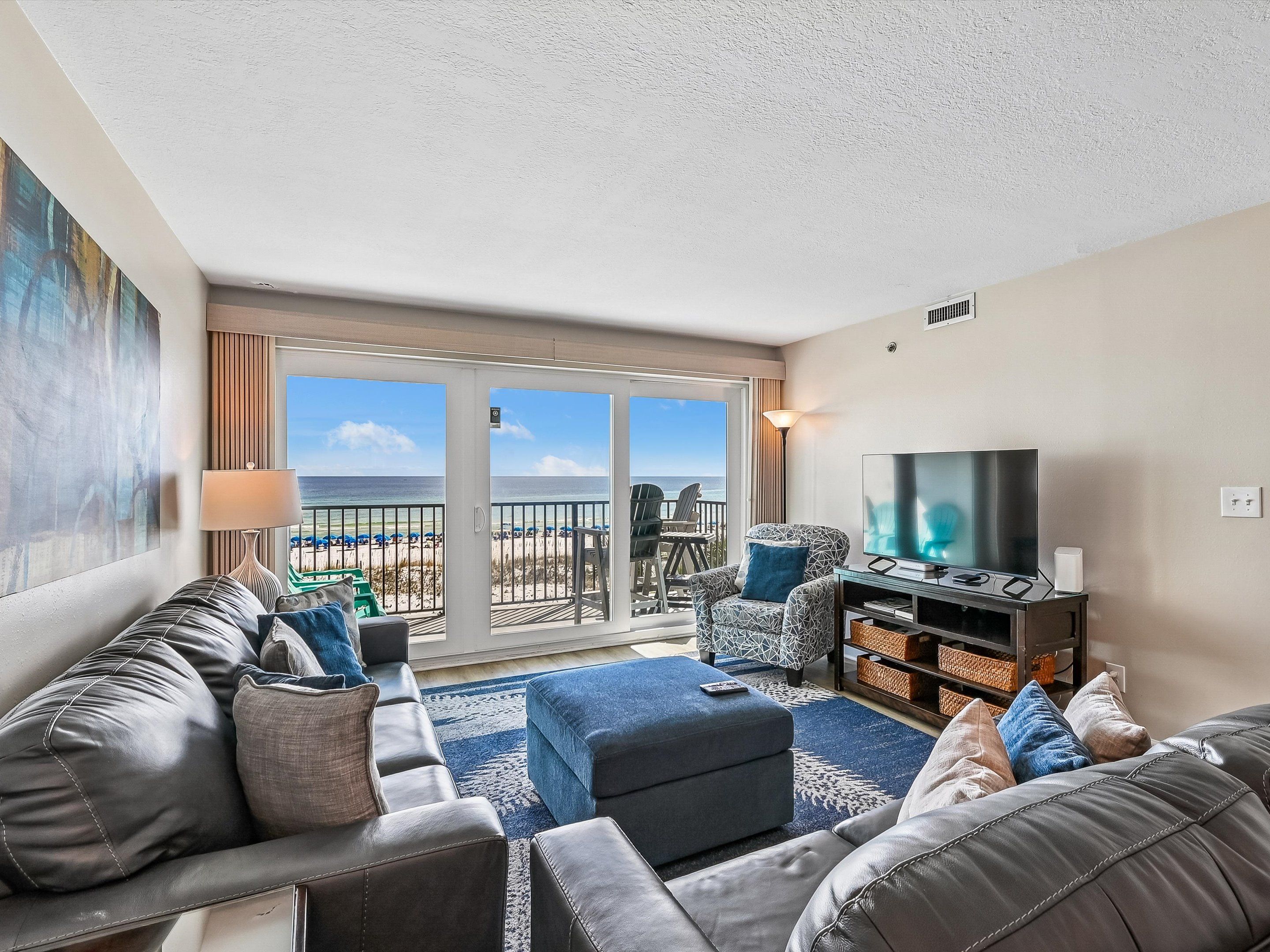 Ein Wohnzimmer mit einer Sofagarnitur, einem Fernseher und Blick auf den Strand durch eine Schiebetür.