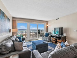 Una sala de estar con un conjunto de sofás, un televisor y vista a la playa a través de un ventanal corredizo.