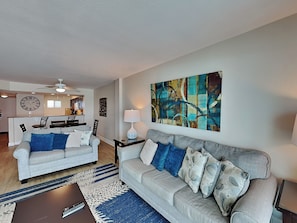 A living room with a sofa, a coffee table, a painting, and a dining area with a clock.