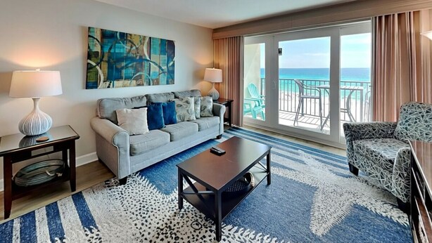 Une chambre d’hôtel avec un canapé, une table basse, un fauteuil, un tableau et un balcon donnant sur l’océan.