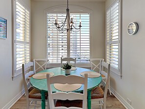 A dining area with a round table, white chairs, a chandelier, and a view through large windows with blinds. - Beach Colony Towers #9D | Gulf Front Condo w. Beach + Pool Access (Perdido Key)
