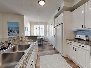 A kitchen with white cabinets, granite countertops, stainless steel sinks, and a refrigerator. - Beach Colony Towers #9D | Gulf Front Condo w. Beach + Pool Access (Perdido Key)