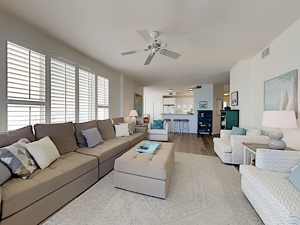 A spacious living room with a large sofa, ottoman, and a dining area with a kitchen in the background. - Beach Colony Towers #9D | Gulf Front Condo w. Beach + Pool Access (Perdido Key)