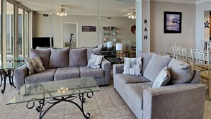 A spacious living room with two grey sofas, a glass coffee table, and a dining area with a white table and chairs. - Water's Edge #305 | Great Location + Pool! (Fort Walton Beach)