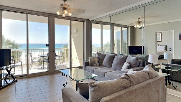 A spacious living room with a large glass door leading to a balcony with a view of the ocean. There is a sofa, a coffee table, and a TV on a stand. - Water's Edge #305 | Great Location + Pool! (Fort Walton Beach)