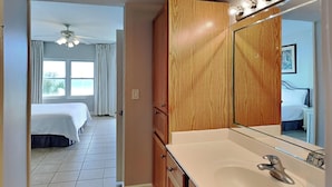 A hotel room with a large wooden wardrobe, a bathroom with a mirror and sink, and a bed with a white bedspread. - Water's Edge #305 | Great Location + Pool! (Fort Walton Beach)