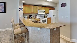 A kitchen with wooden cabinets, a built-in island with a sink, a microwave, a stove, and a refrigerator. - Water's Edge #305 | Great Location + Pool! (Fort Walton Beach)