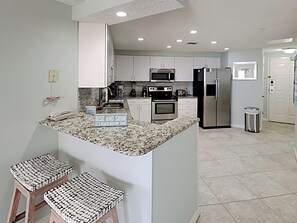 A modern kitchen with white cabinets, granite countertops, and stainless steel appliances. There are two bar stools with woven seats.