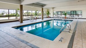 En indendørs swimmingpool med krystalklart vand, omgivet af liggestole og potteplanter.