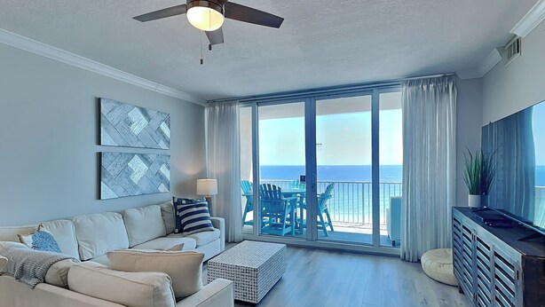 A spacious living room with a ceiling fan, a large window with a view of the ocean, a sofa with cushions, and a TV on a wooden cabinet. - Gulf Front Condo with Stunning View and Resort Amenities (Gulf Shores)