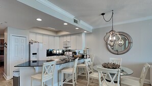 A modern kitchen with white cabinetry, a central island with bar stools, and a dining area with a round table and chairs. - Gulf Front Condo with Stunning View and Resort Amenities (Gulf Shores)