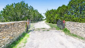A gravel road leading through a stone wall gate surrounded by dense green trees. - 60-Acre Retreat w/ Hot Tub, Firepit & Pet-Friendly! (Wimberley)