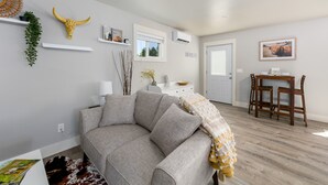 Un salon moderne avec un canapé d’angle gris, un coin repas avec une table et des chaises en bois, et des éléments de décoration murale, notamment un crâne de taureau jaune et un tableau encadré représentant une vache.