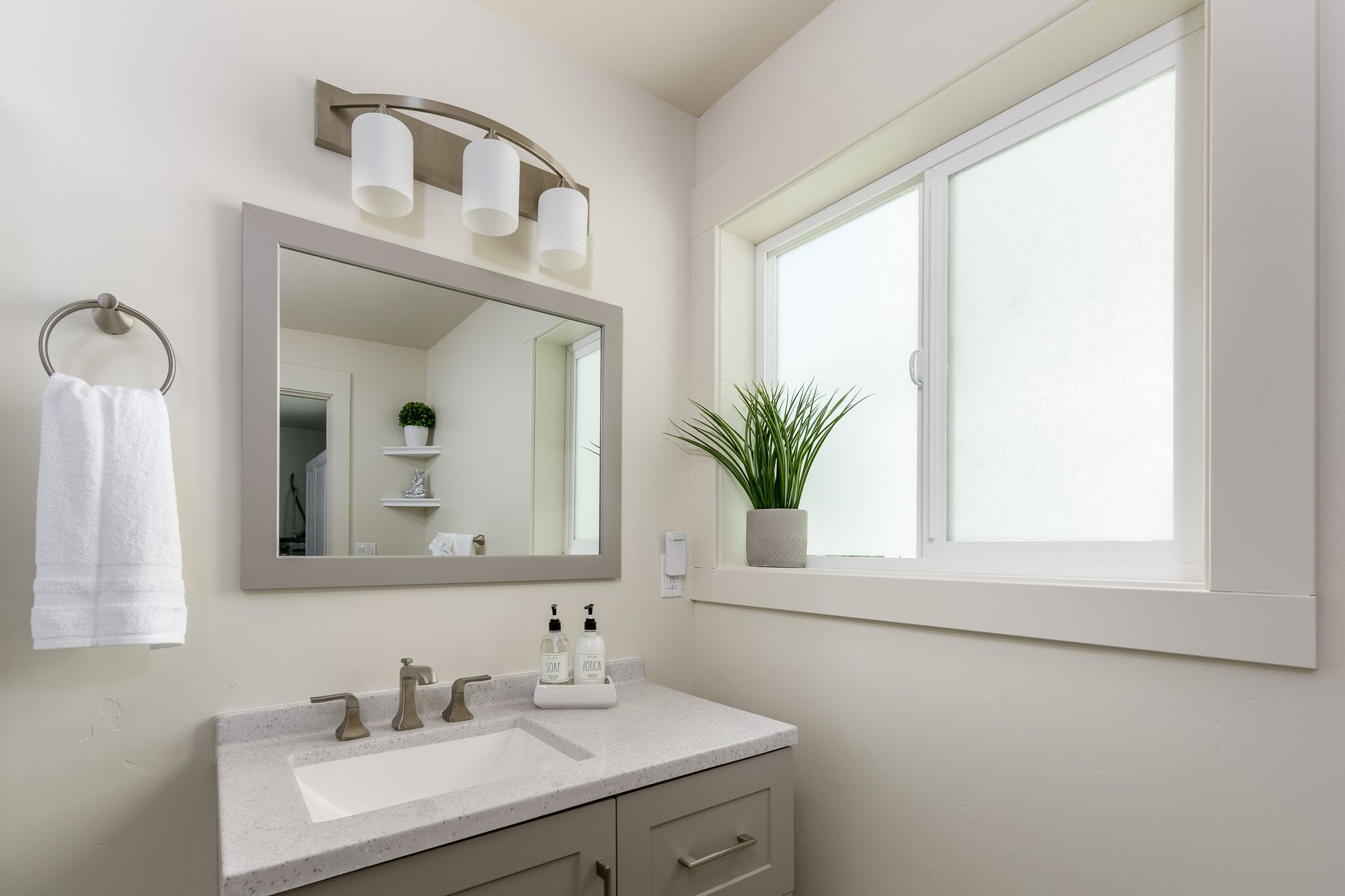 Une salle de bain avec un grand miroir, un lavabo à deux robinets, un porte-serviettes et une fenêtre avec une plante.