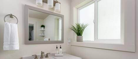 Une salle de bain avec un grand miroir, un lavabo à deux robinets, un porte-serviettes et une fenêtre avec une plante.