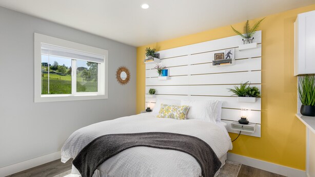 Une chambre à coucher comprenant un lit, une table de chevet, une commode, une étagère murale ornée de plantes, une fenêtre et une vue sur la verdure à l’extérieur.