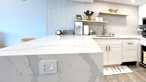 A modern kitchen with a marble countertop, white cabinets, and a built-in sink. There is a clock on the wall and a shelf with decorative items. - New Aloha Waikiki Ilikai Hotel Condo Free Parking (Honolulu)