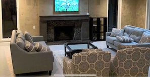 Cozy living room with a fireplace, two sofas, and a coffee table, featuring a large window, bookshelf, and mounted television.
