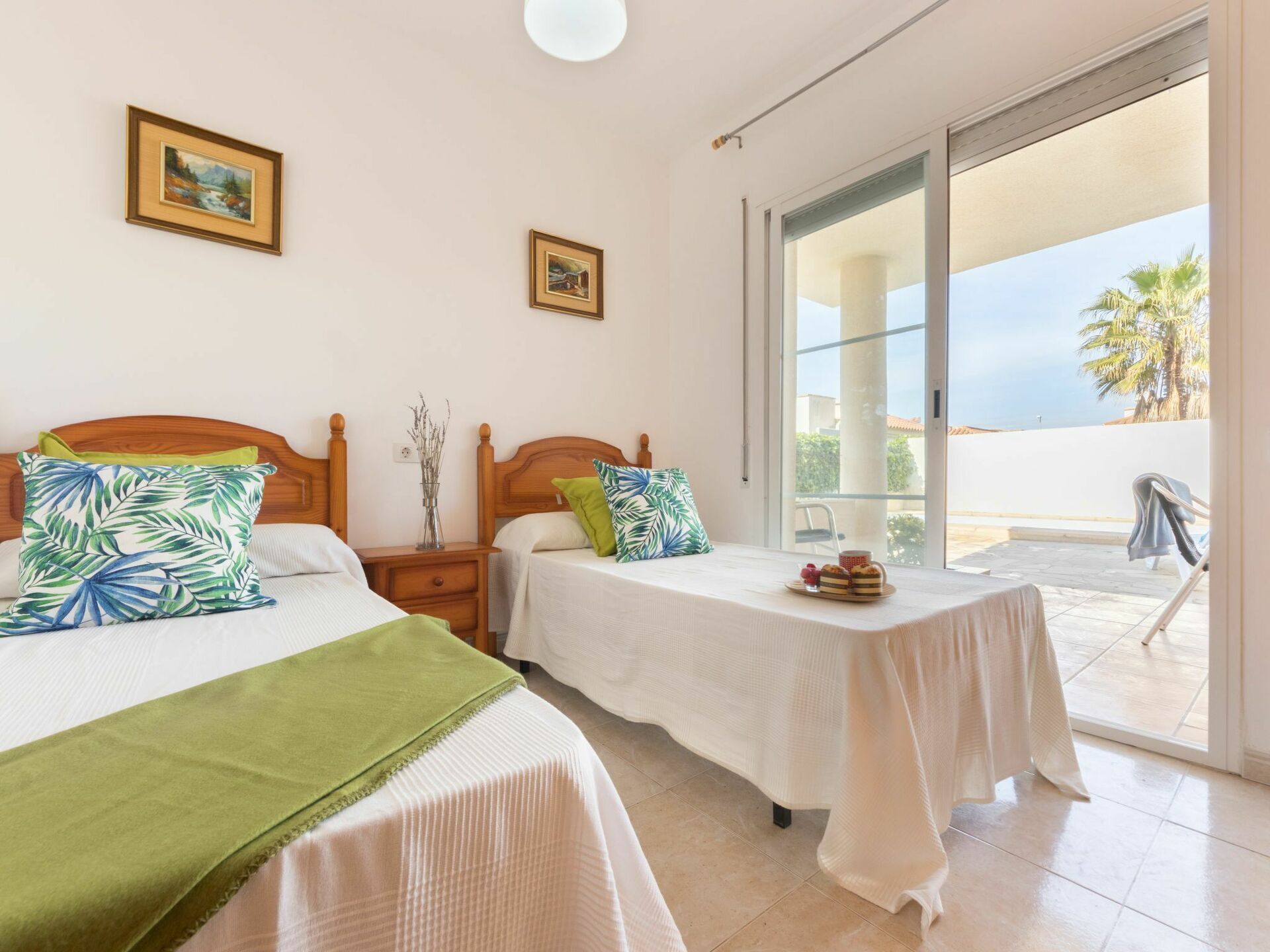 A hotel room with two beds, a balcony with a view of palm trees, and a table with a plate of pastries.