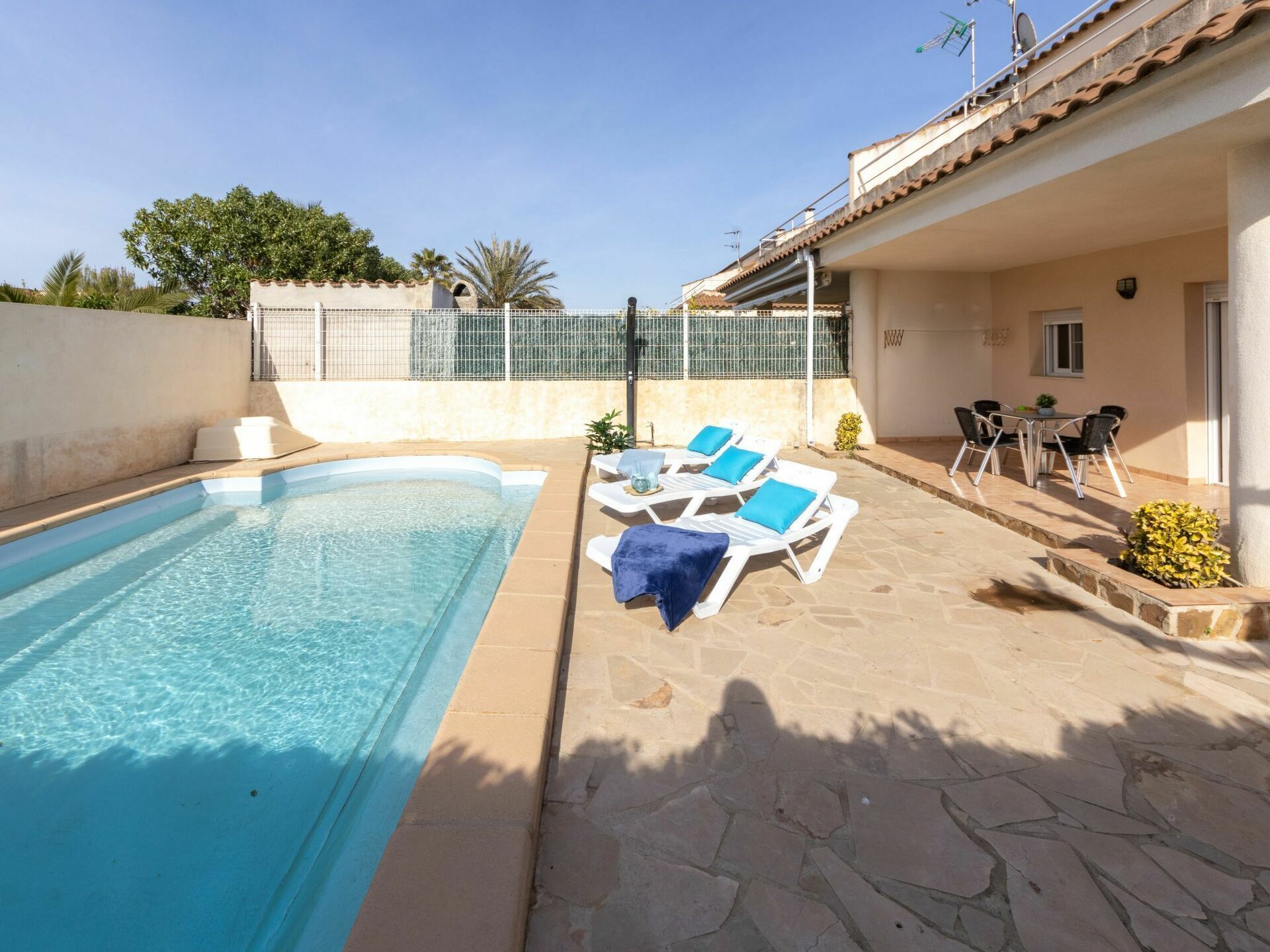A swimming pool with lounge chairs and a table, surrounded by stone flooring and a covered patio area.