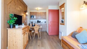 A compact kitchen with wooden cabinets, a dining table, and chairs. A red door leads to another room.