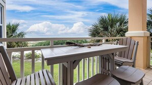 A balcony with a table and chairs, overlooking a grassy area and palm trees under a clear sky. - PERFECT FORT MORGAN GETAWAY, ROOM FOR THE WHOLE FAMILY TO ENJOY! (Gulf Shores)