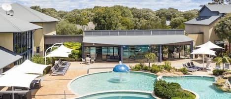 Une piscine avec des chaises longues, un espace de détente couvert et un bâtiment doté de grandes fenêtres.