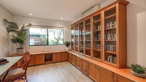 A room with a large wooden cabinet with glass doors, a dining table with chairs, and a window with a view of greenery outside. - Baydream Believer: Lush Beachfront Resort Escape (Busselton)