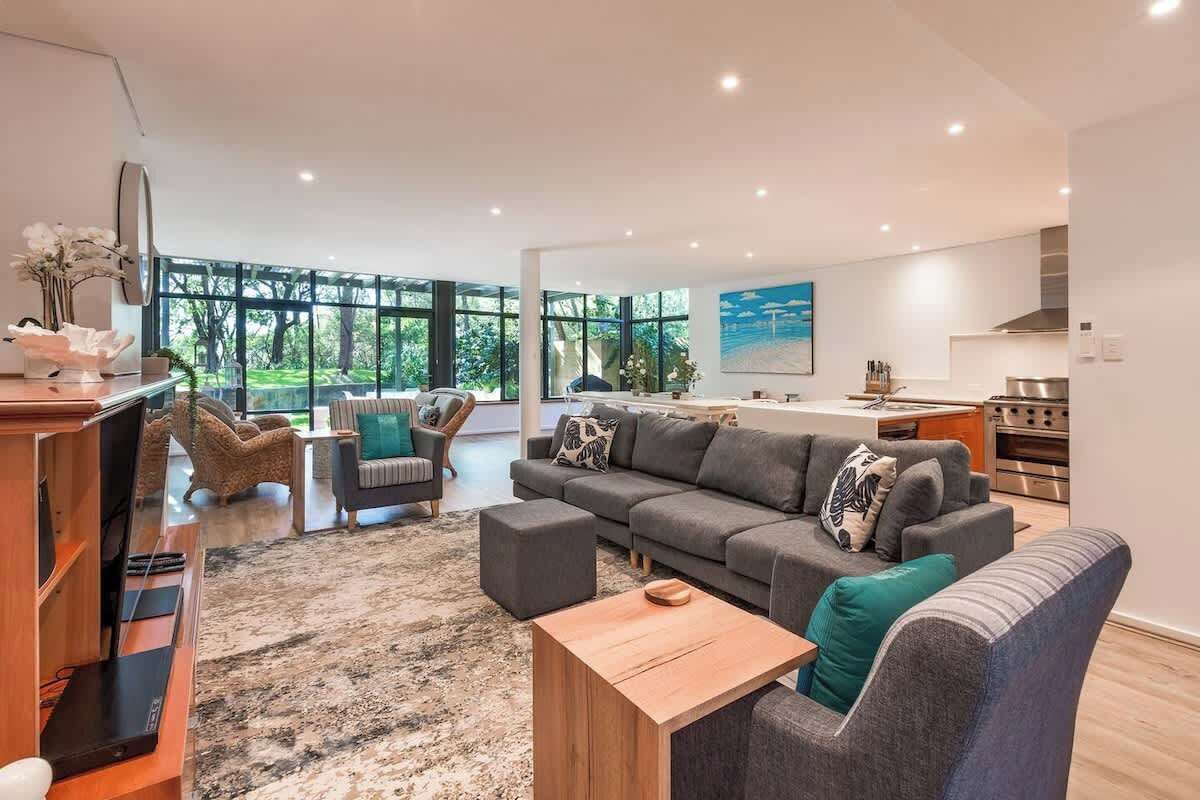 Un salon moderne avec un grand canapé d’angle gris, une table basse en bois et un téléviseur posé sur un meuble en bois. On y trouve également un coin cuisine avec une cuisinière et un four, ainsi qu’une table à manger avec des chaises.