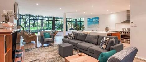Un salon moderne avec un grand canapé d’angle gris, une table basse en bois et un téléviseur posé sur un meuble en bois. On y trouve également un coin cuisine avec une cuisinière et un four, ainsi qu’une table à manger avec des chaises.
