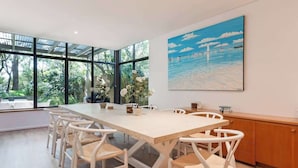 A modern dining area with a long table and white chairs, large windows, and a painting of a seascape. - Baydream Believer: Lush Beachfront Resort Escape (Busselton)