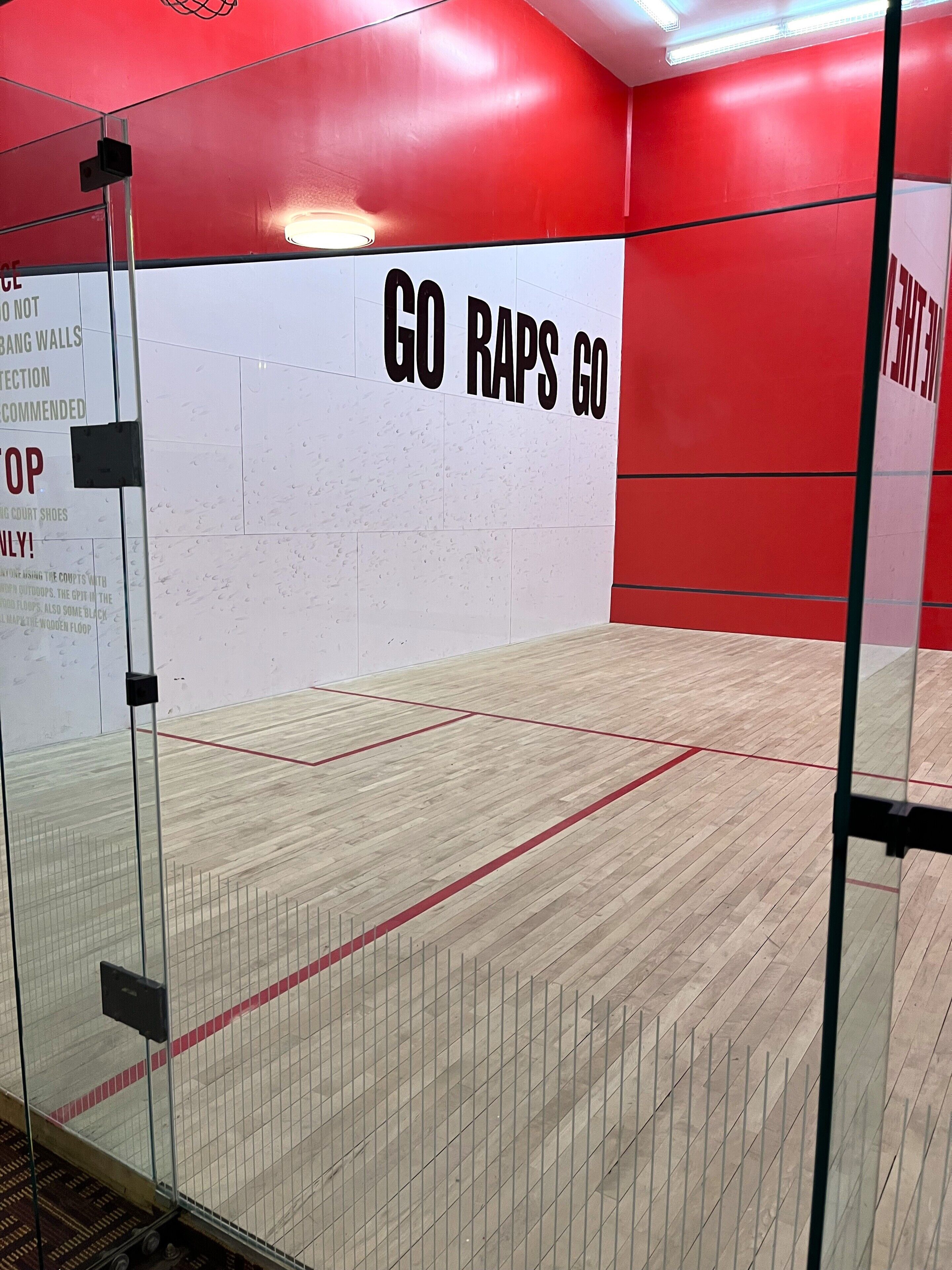 A squash court with a red and white wall, a wooden floor, and a glass door.