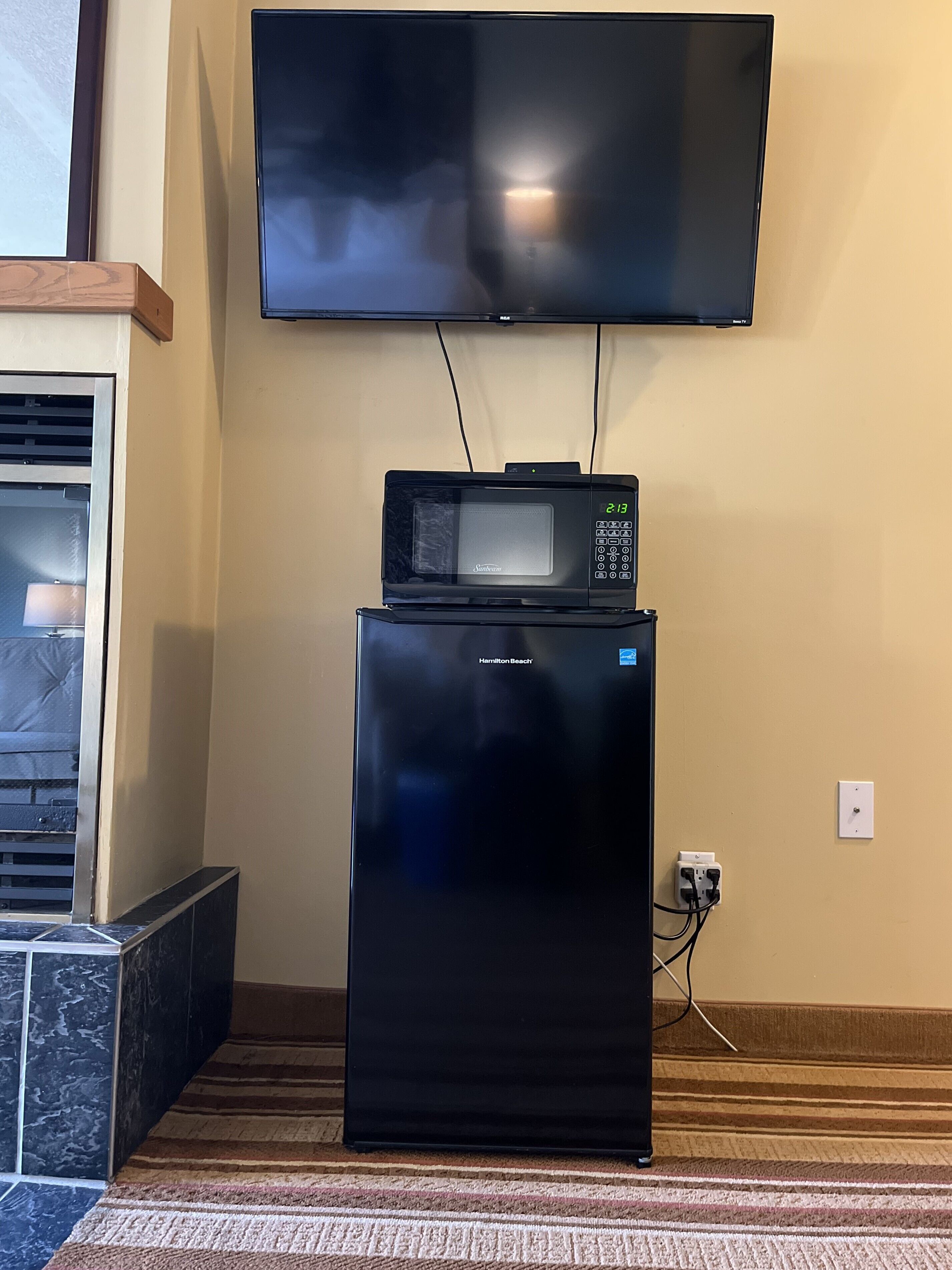 A black refrigerator with a microwave on top, a flat-screen TV mounted on the wall, and a power outlet on the wall.