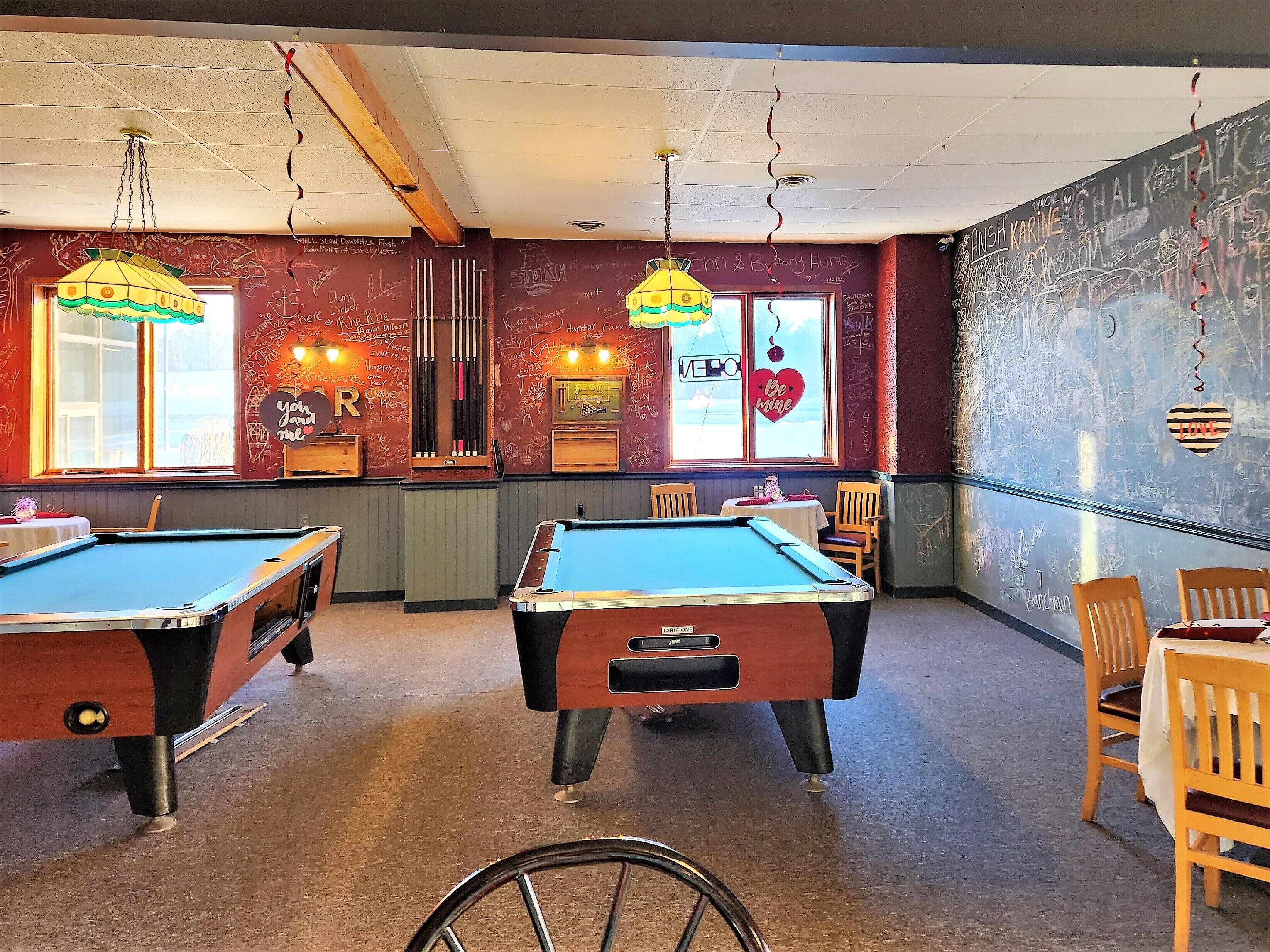 A pool hall with two pool tables, wooden chairs, and a bicycle in the foreground.