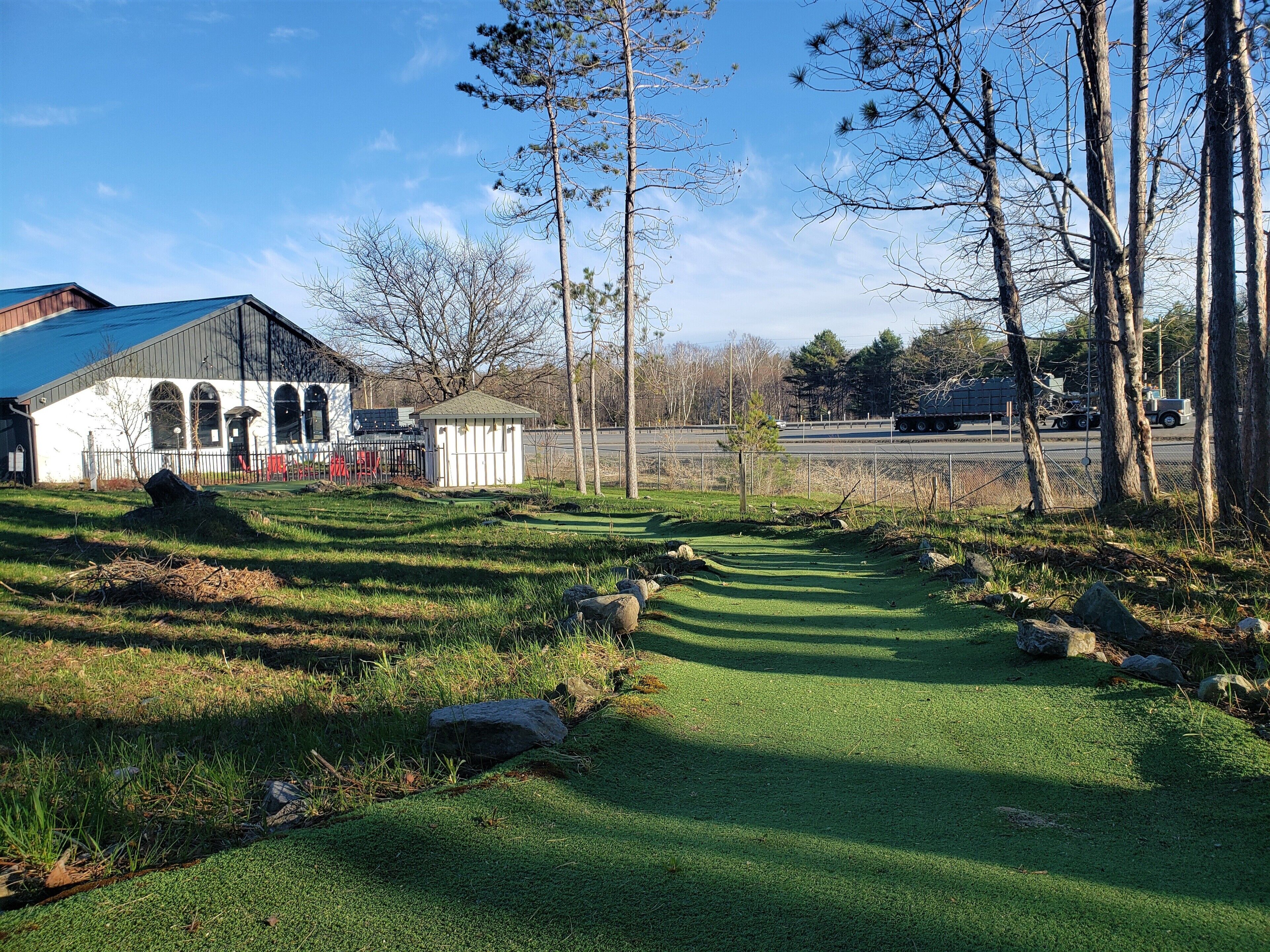 A golf course with a clubhouse, a small pavilion, and a road with vehicles in the background.