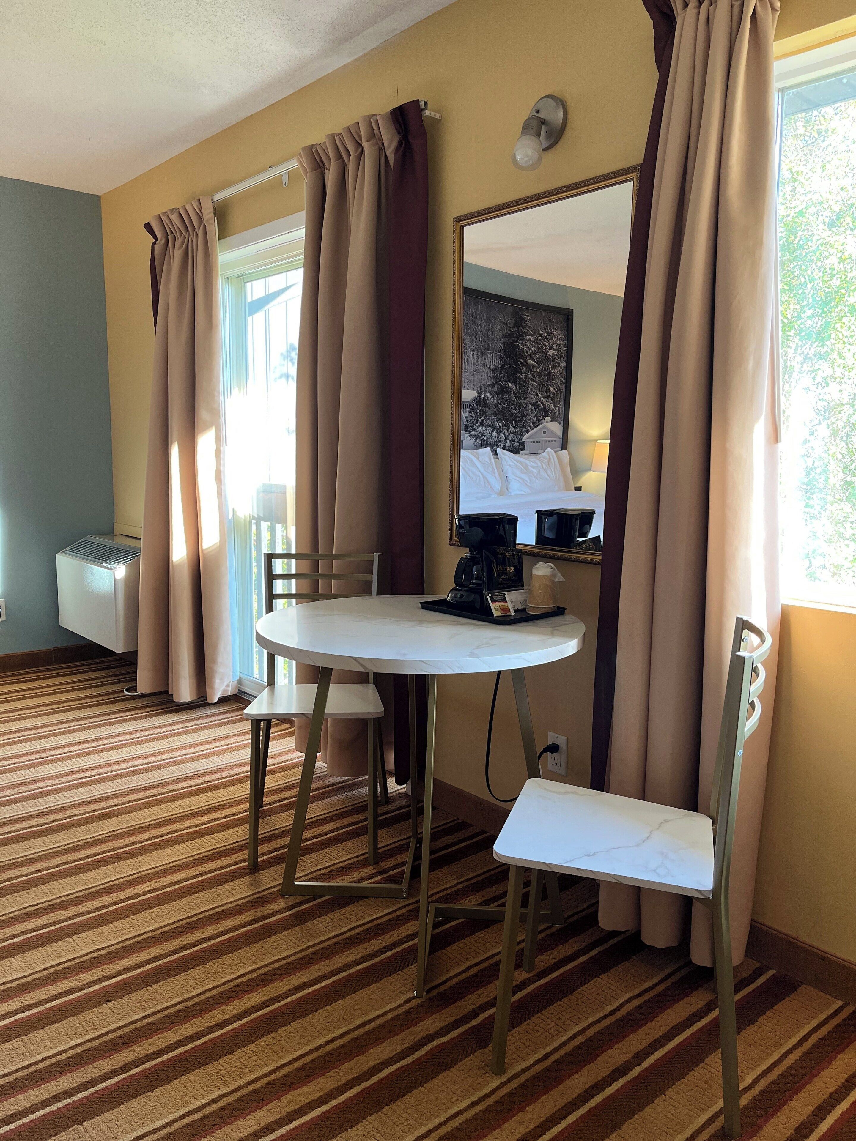 A hotel room with a round table, two chairs, a large mirror, and a window with curtains.
