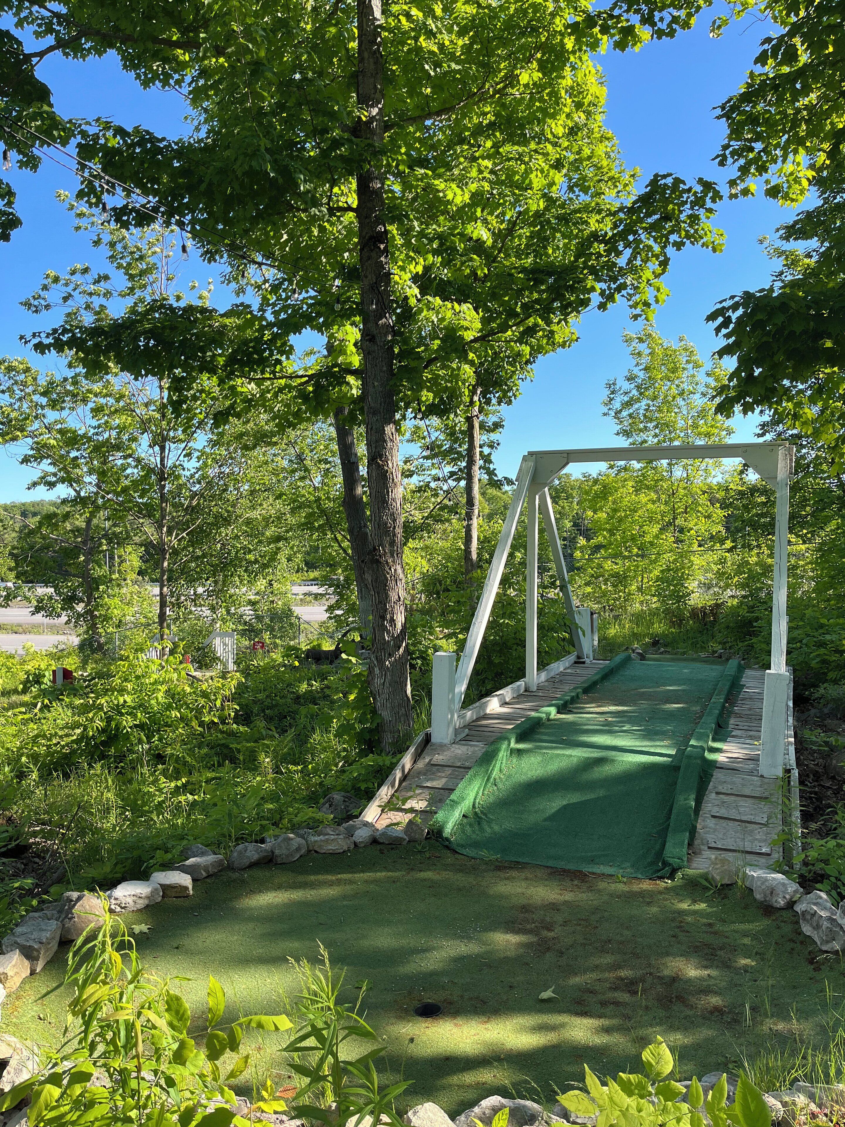 Un putting green de césped artificial verde, con una estructura de puente de madera blanca, rodeado de exuberante follaje y árboles.