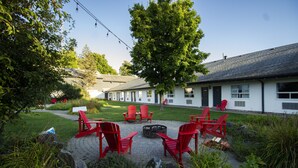 A courtyard with red Adirondack chairs arranged around a fire pit, a white building with multiple windows, and a tree providing shade. - Jolly Roger Inn & Resort (Seguin)