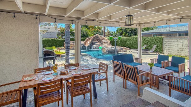 A covered patio with a pool, wooden furniture, and a view of a landscaped garden. - Walk to Disney + Brand New Pool and Rockslide + Game Room + Pet Friendly (Anaheim)