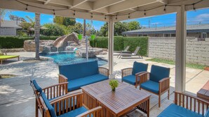 A covered patio area with a pool, lounge chairs, and a wooden table with blue cushions. - Walk to Disney + Brand New Pool and Rockslide + Game Room + Pet Friendly (Anaheim)