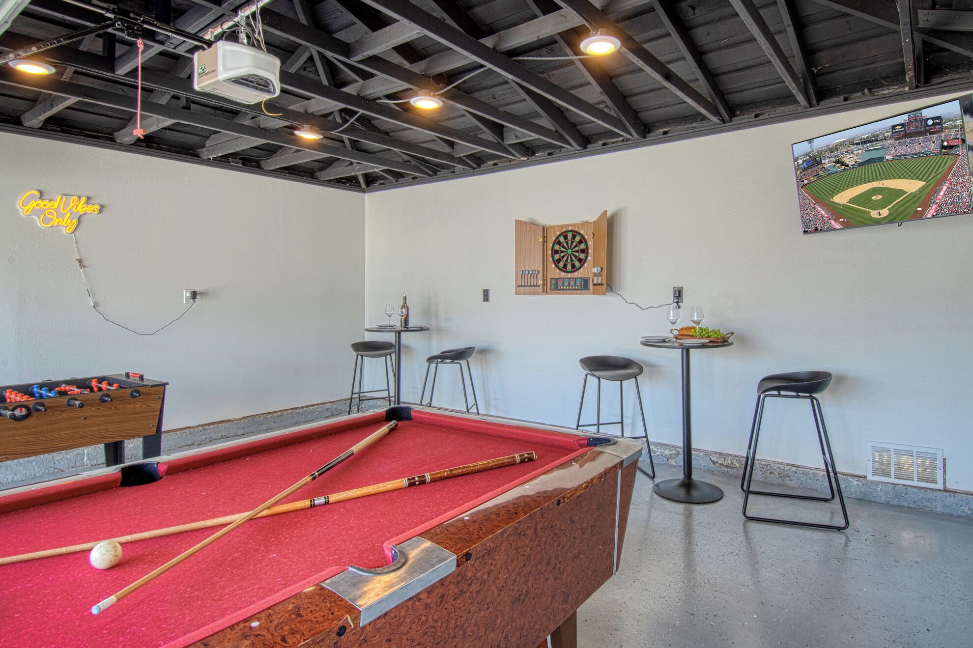 A billiards table with pool cues, a dartboard, and a small table with wine glasses and a fruit bowl.