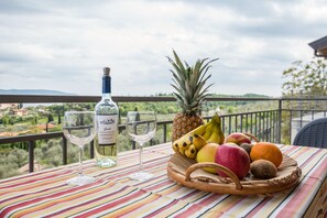 Un tavolo con una bottiglia di vino, due bicchieri e un cesto di frutta su un balcone affacciato su vegetazione ed edifici.