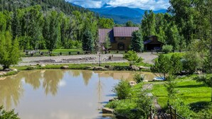 A rustic lodge with a metal roof, surrounded by a pond, trees, and mountains in the background. - Riverfront, Hot Tub, Pond, Fire Pit, Stables, Deck, Near Downtown Pagosa! (Pagosa Springs)