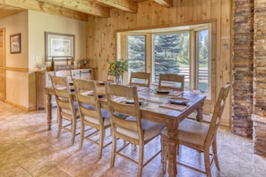 Une salle à manger en bois, avec une grande table dressée pour six personnes, bordée de murs en pierre et dotée de grandes fenêtres offrant une vue sur l’extérieur.
