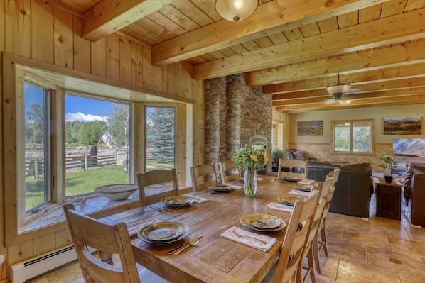 Une salle à manger en bois, avec une grande table dressée pour six personnes, une cheminée en pierre et une vue sur l’extérieur.