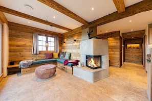 A log cabin interior with a stone fireplace, a seating area with cushions, and a large window. - Alm Lenze Chalet, 130 m², sleeps 4, wood-burning stove (Samerberg)
