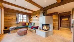 Un acogedor interior de cabaña de madera con chimenea de piedra, vigas de madera y una zona de estar con cojines.